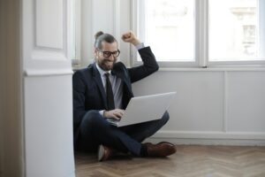 pexels-photo-3785263-3785263 Cheerful man sitting on the floor with a laptop, celebrating work success.